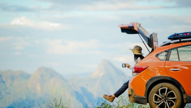 A woman sits on the tailgate of an orange Subaru drinking coffee out of a metal cup while parked at a scenic mountain vista