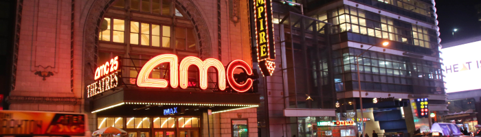 Wide, short photo of an AMC theater building in a city at night with the AC logo visible near the center